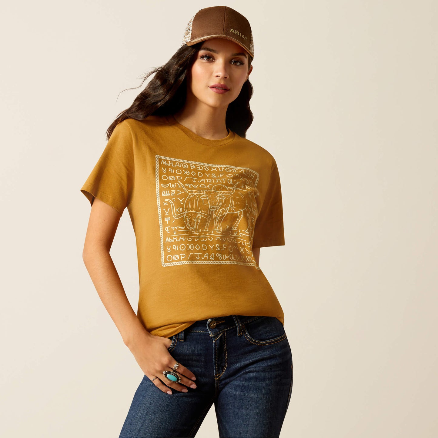 Woman wearing a mustard yellow t-shirt with a graphic design, blue jeans, and a brown cap on a beige background