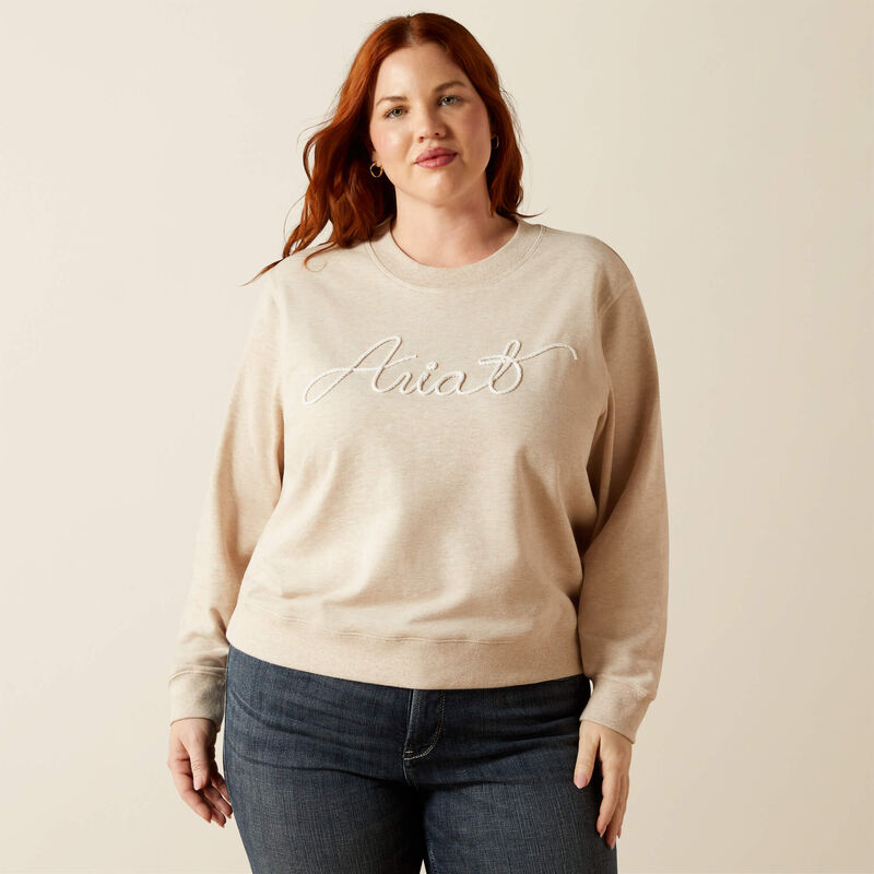 Plus size woman wearing beige Ariat logo sweatshirt and dark jeans in neutral setting