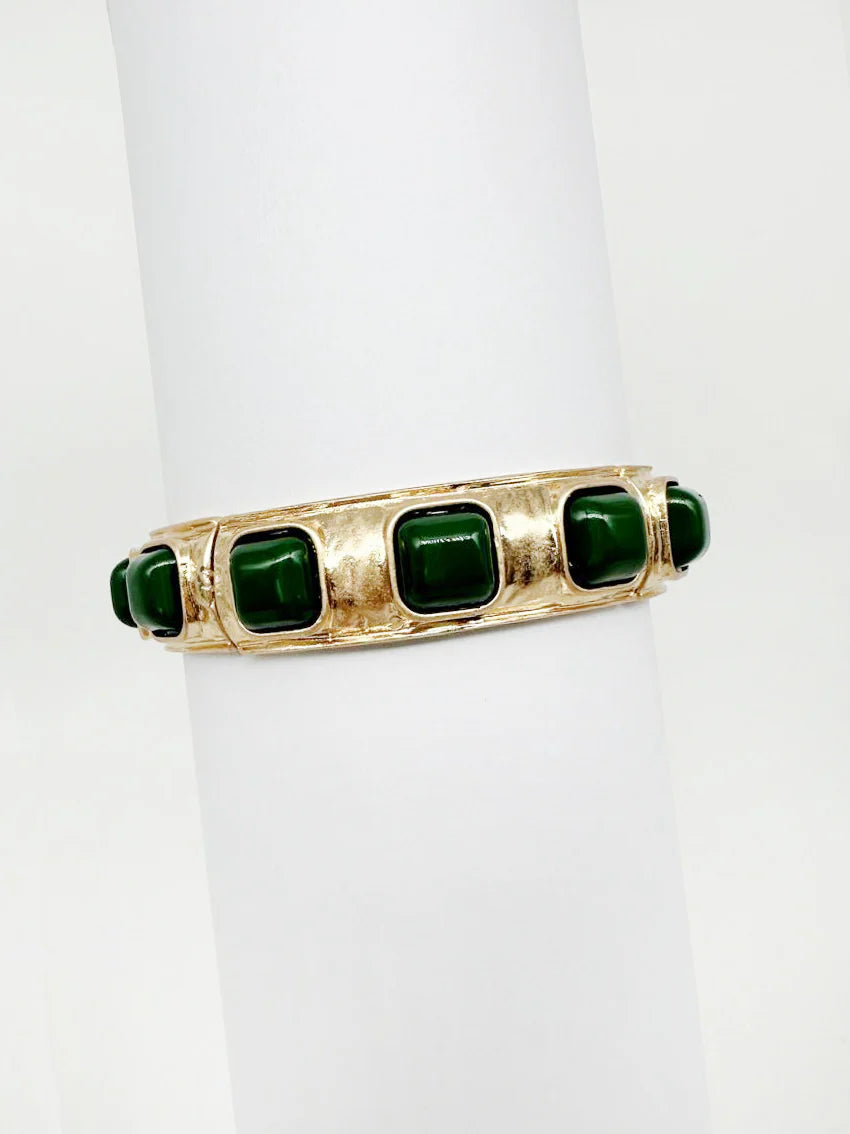 Gold cuff bracelet with square green stones, displayed on a white cylinder