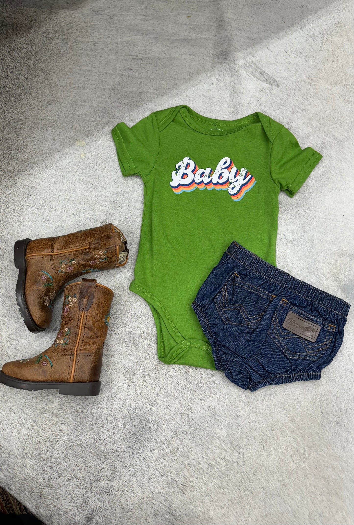 Green baby onesie with 'Baby' print, denim shorts, and brown embroidered boots on fur background