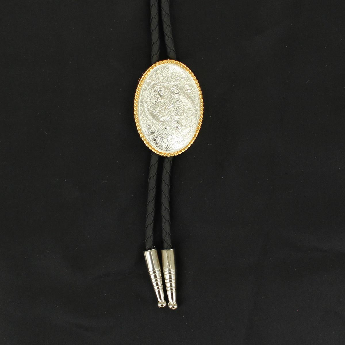 A western-style bolo tie featuring a black braided cord, a silver-toned engraved oval concho with a gold-toned border, and silver-toned metal tips, displayed on a black fabric background.