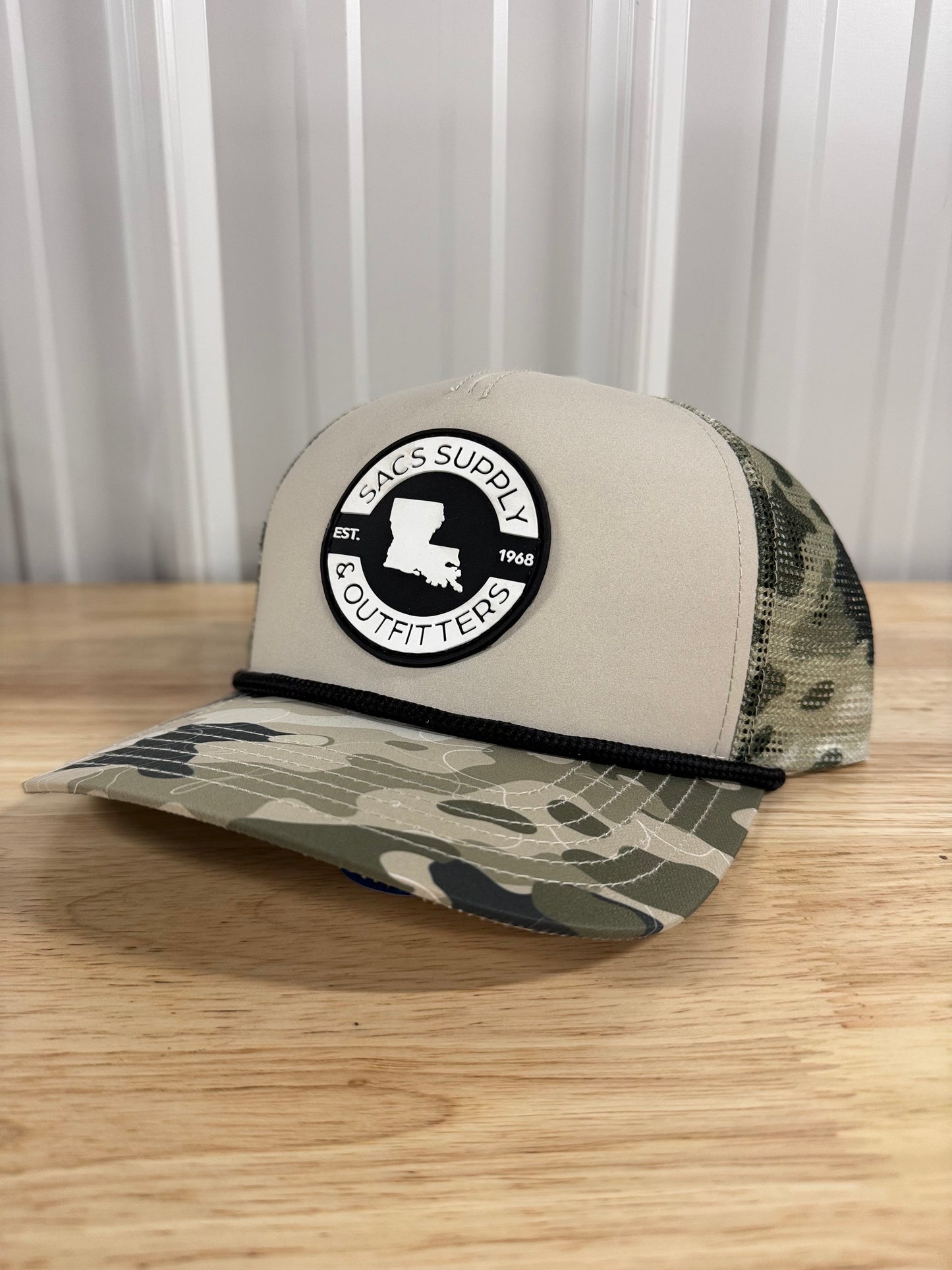 Camo trucker hat with mesh back and Sacs Supply & Outfitters patch on wooden table