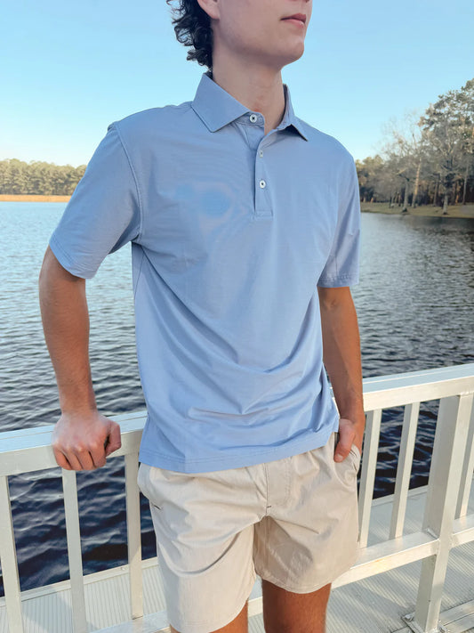 Person wearing a light blue polo shirt and beige shorts standing by a body of water.