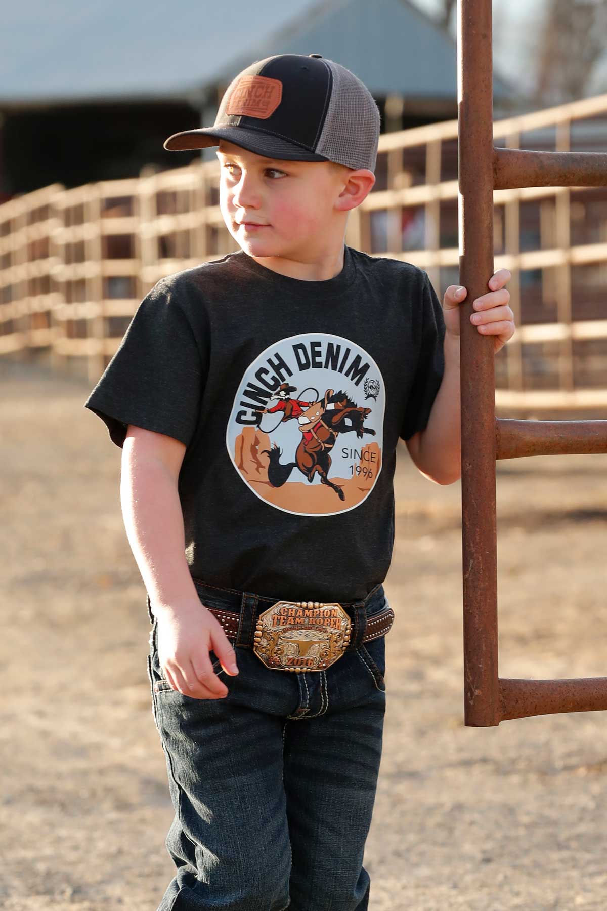 Young boy in black cowboy hat, Cinch Denim graphic t-shirt, jeans, and rodeo belt buckle outdoors