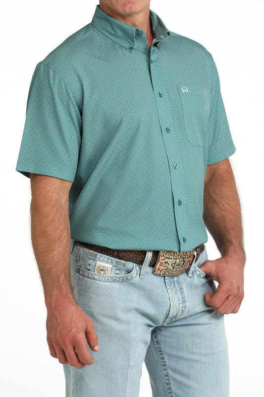 Man wearing a teal patterned button-up shirt and light blue jeans on a white background