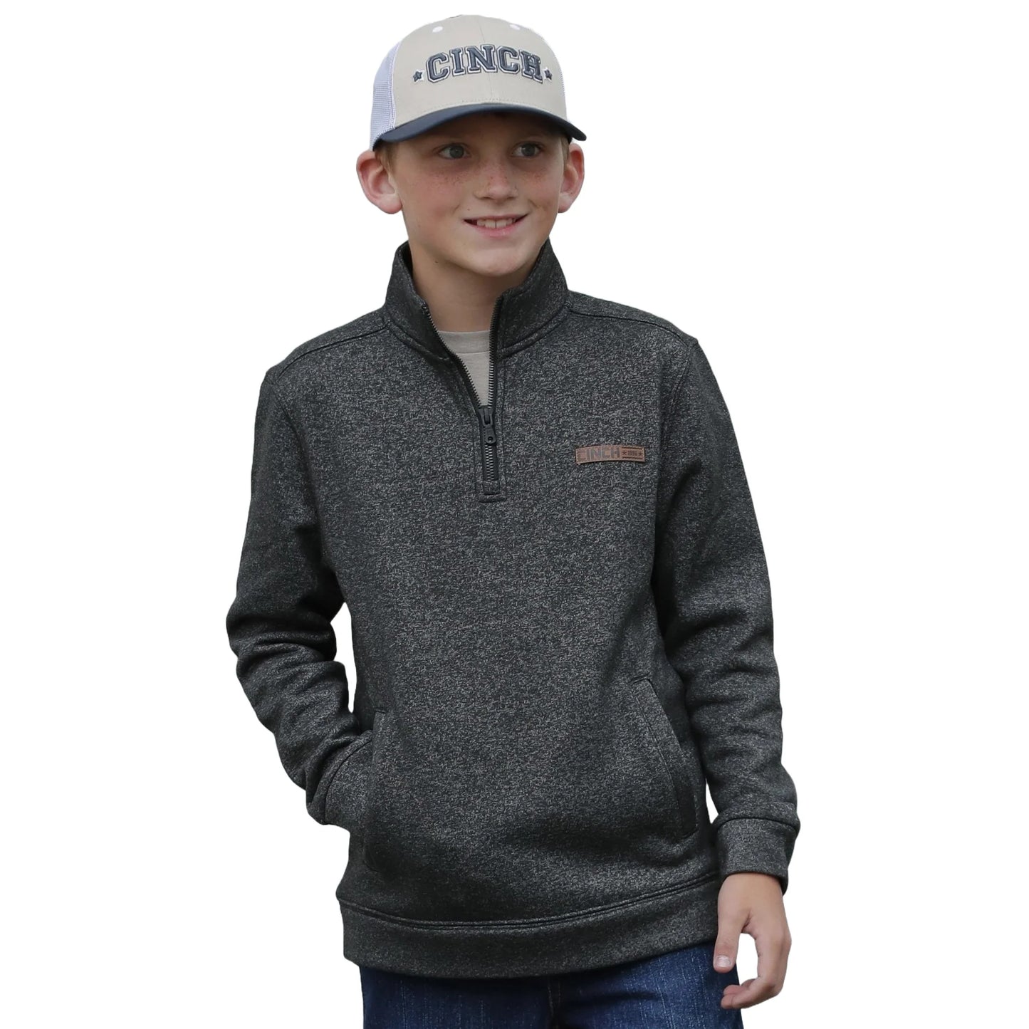 Boy wearing a grey quarter-zip pullover and blue trucker hat with hands in pockets
