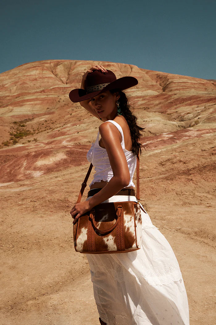 Woman in white dress and cowboy hat with brown cowhide leather shoulder bag in desert landscape