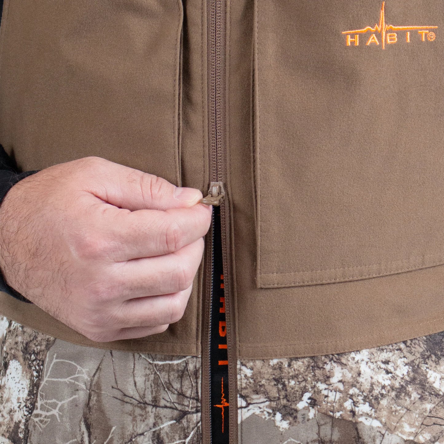 Hand zipping up brown hunting vest with HABIT logo and camo pants