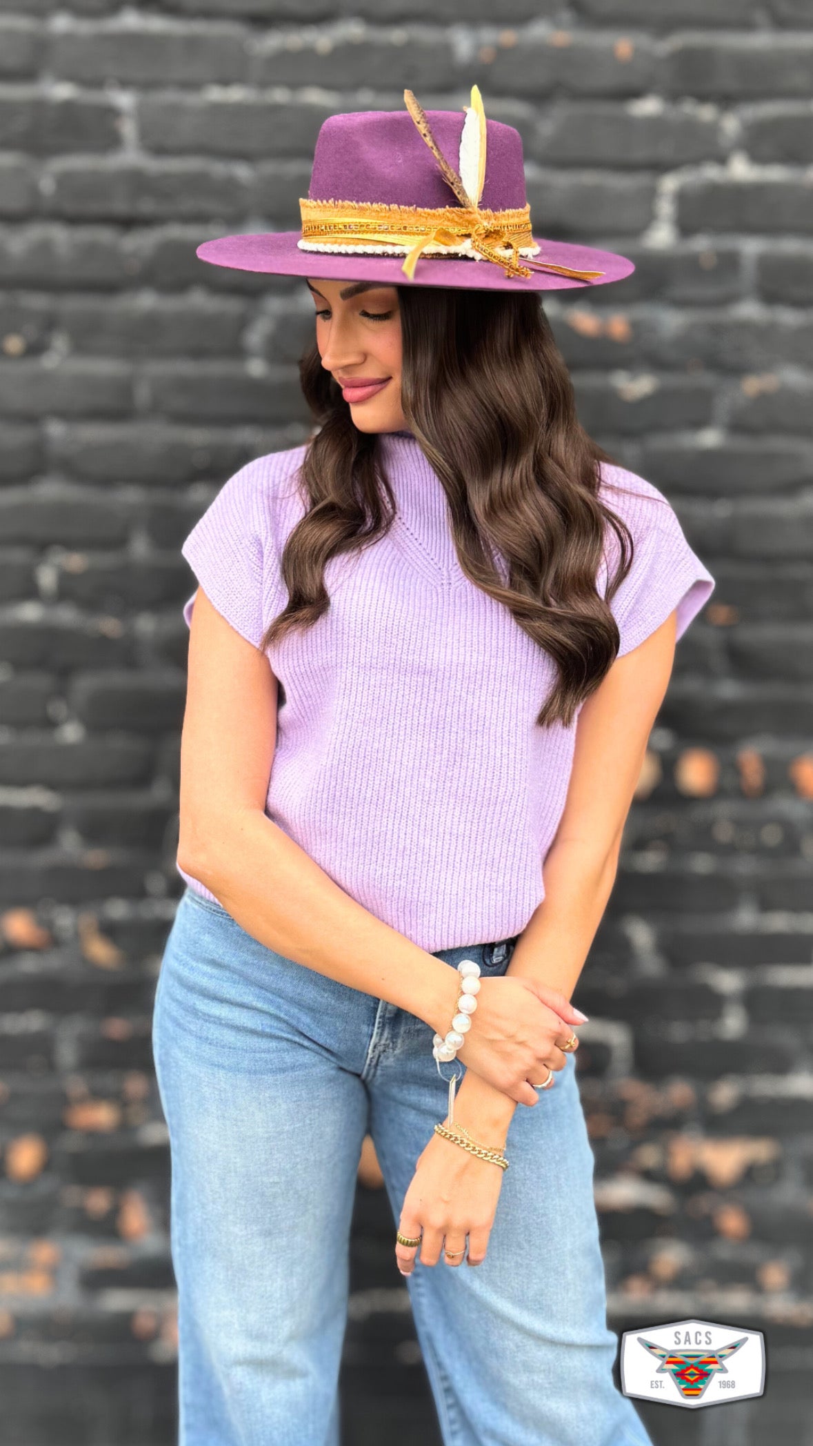 Woman in a purple hat with feather, lilac knit top, and blue jeans posing by brick wall