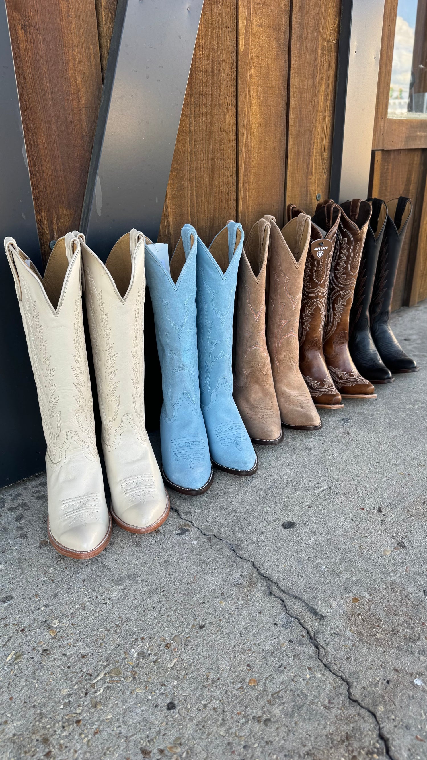 Assorted cowboy boots in various colors lined up against a wooden wall outdoors