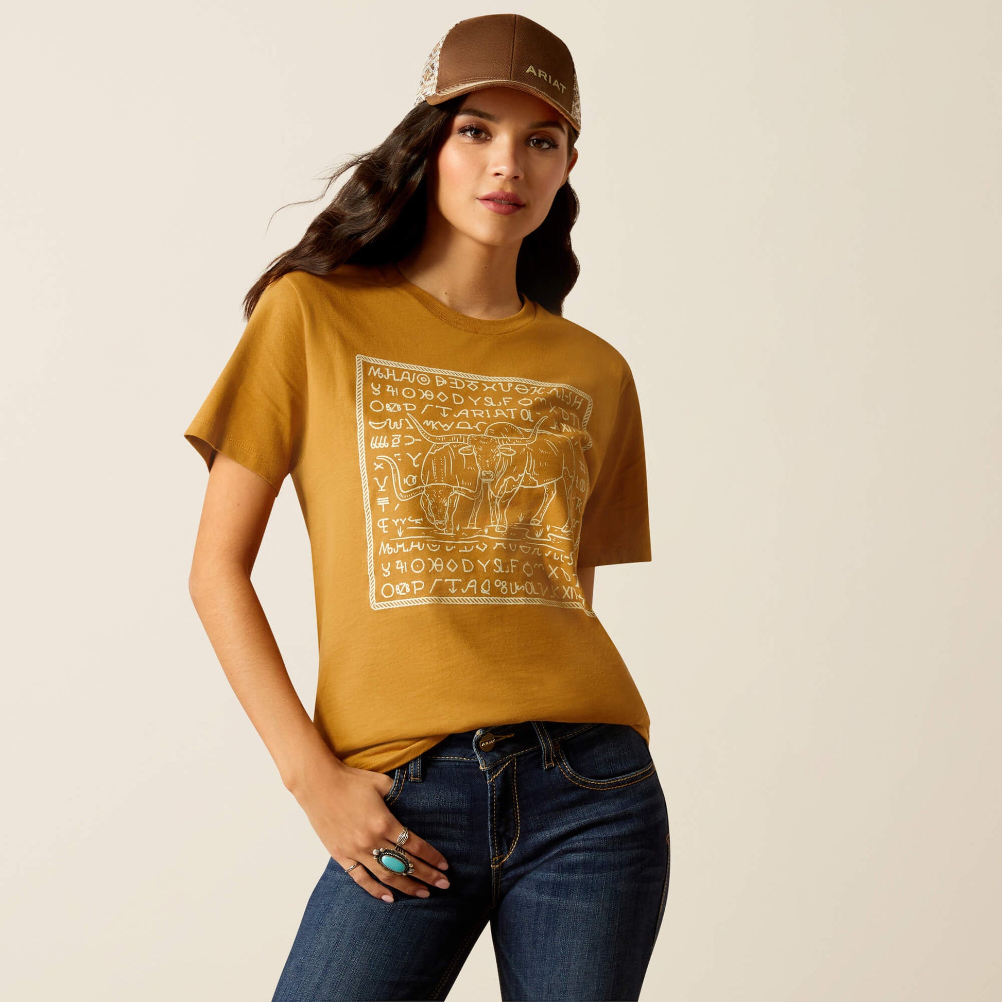 Woman wearing a mustard yellow t-shirt with a graphic design, blue jeans, and a brown cap on a beige background