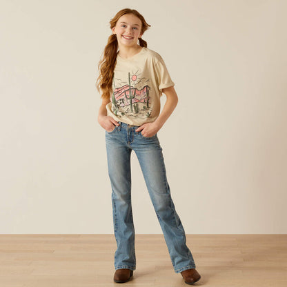 Smiling girl in casual jeans, boots, and a desert-print graphic t-shirt standing indoors