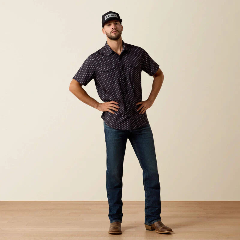 Man wearing a patterned shirt and jeans standing against a plain background
