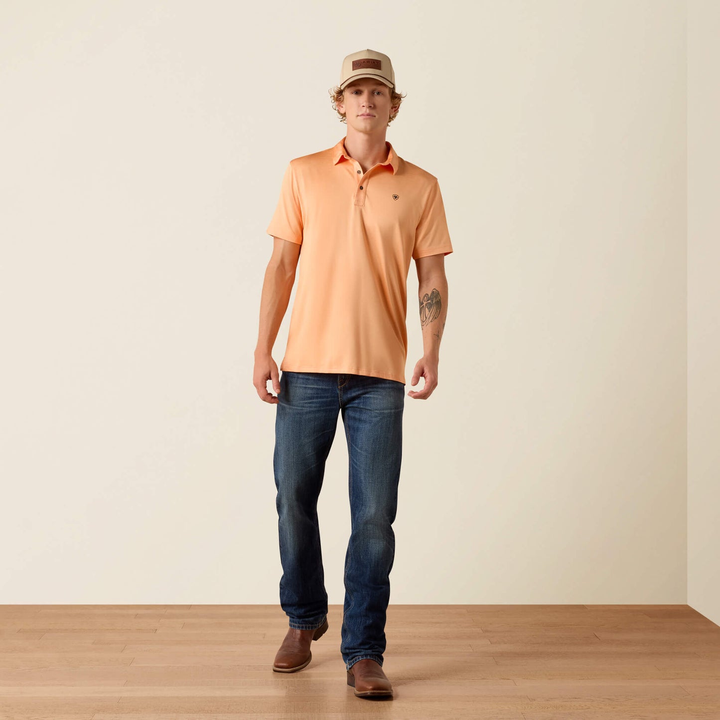 Man wearing an orange polo shirt, blue jeans, and a cap on a plain background