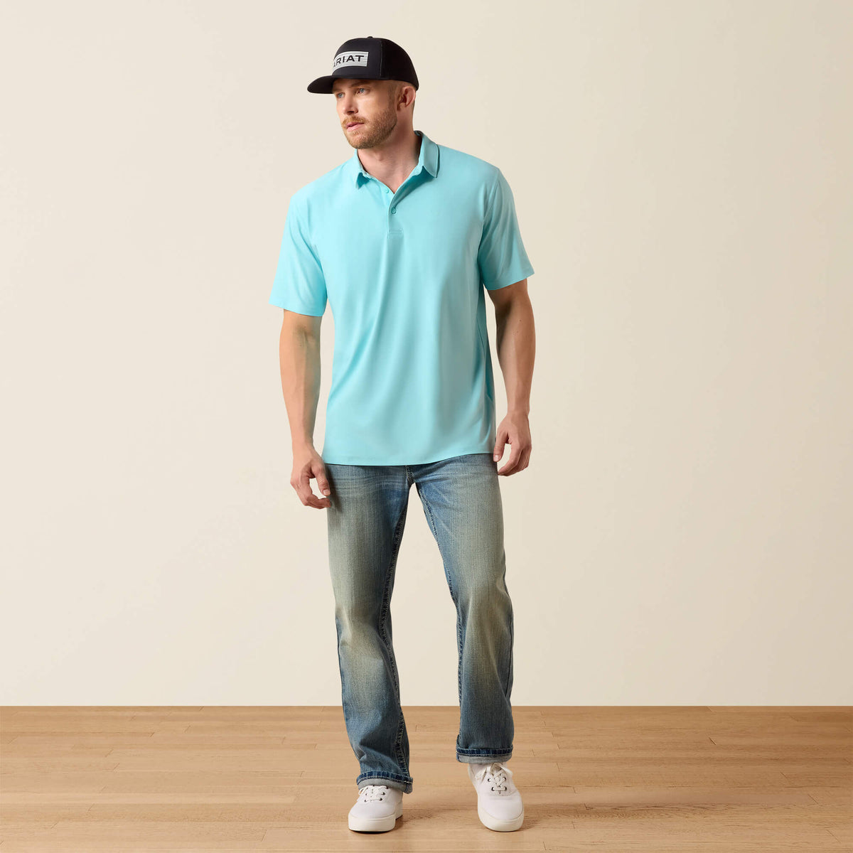 Man wearing a light blue polo shirt, dark jeans, and a black cap with a logo on a beige background