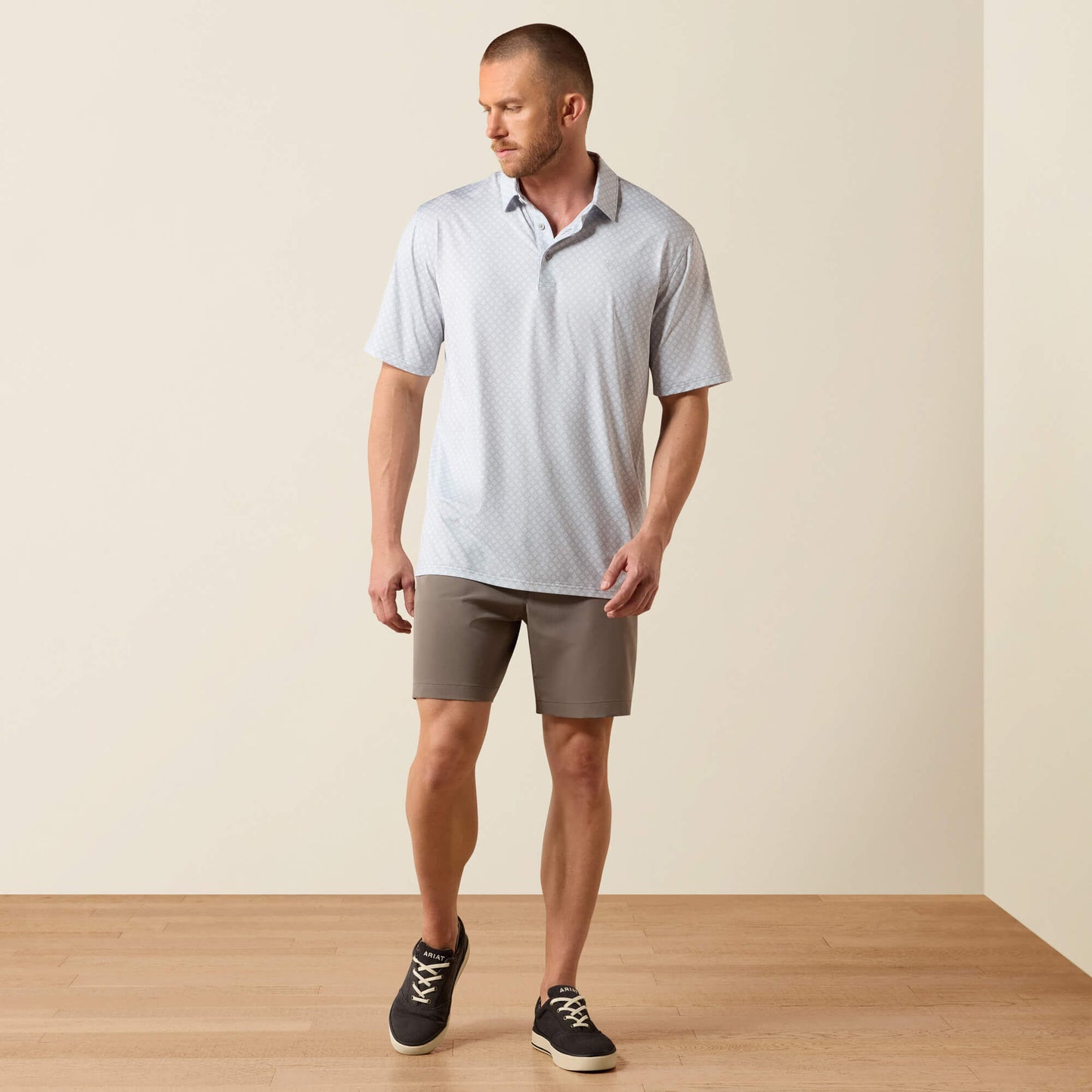 Man wearing a light gray polo shirt and beige shorts standing on a wooden floor with a beige wall background.