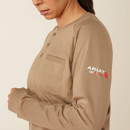 A close-up of a woman wearing a tan long-sleeve henley shirt with a chest pocket and a white and red 'ARIAT CAT' logo on the upper left sleeve, against a plain light beige background.