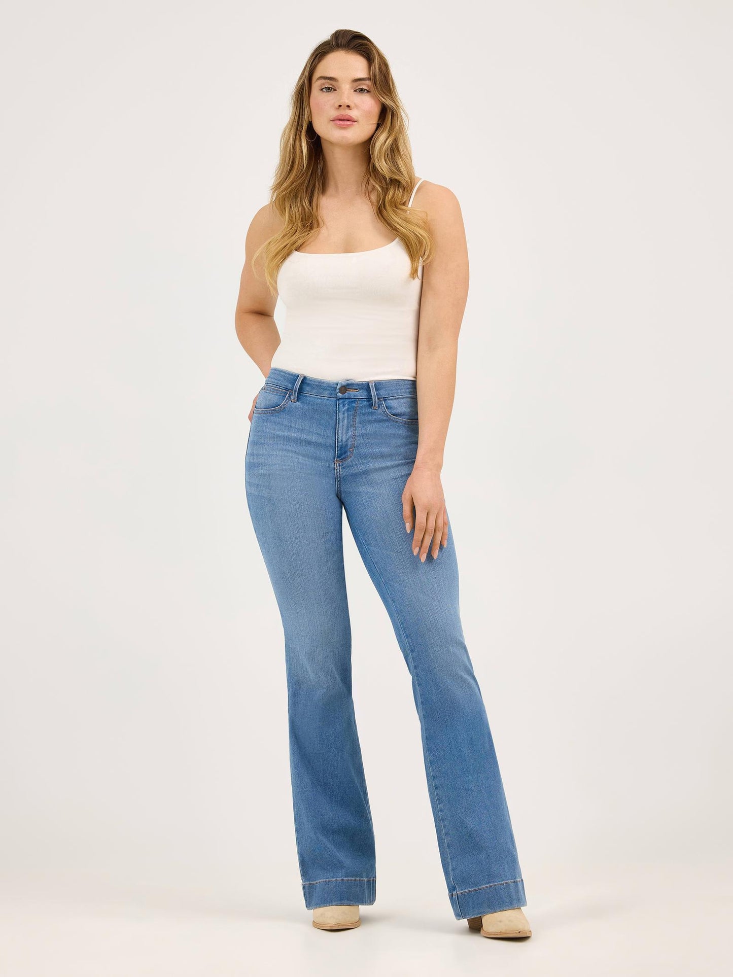 Woman wearing light blue flared jeans and white tank top on a plain background