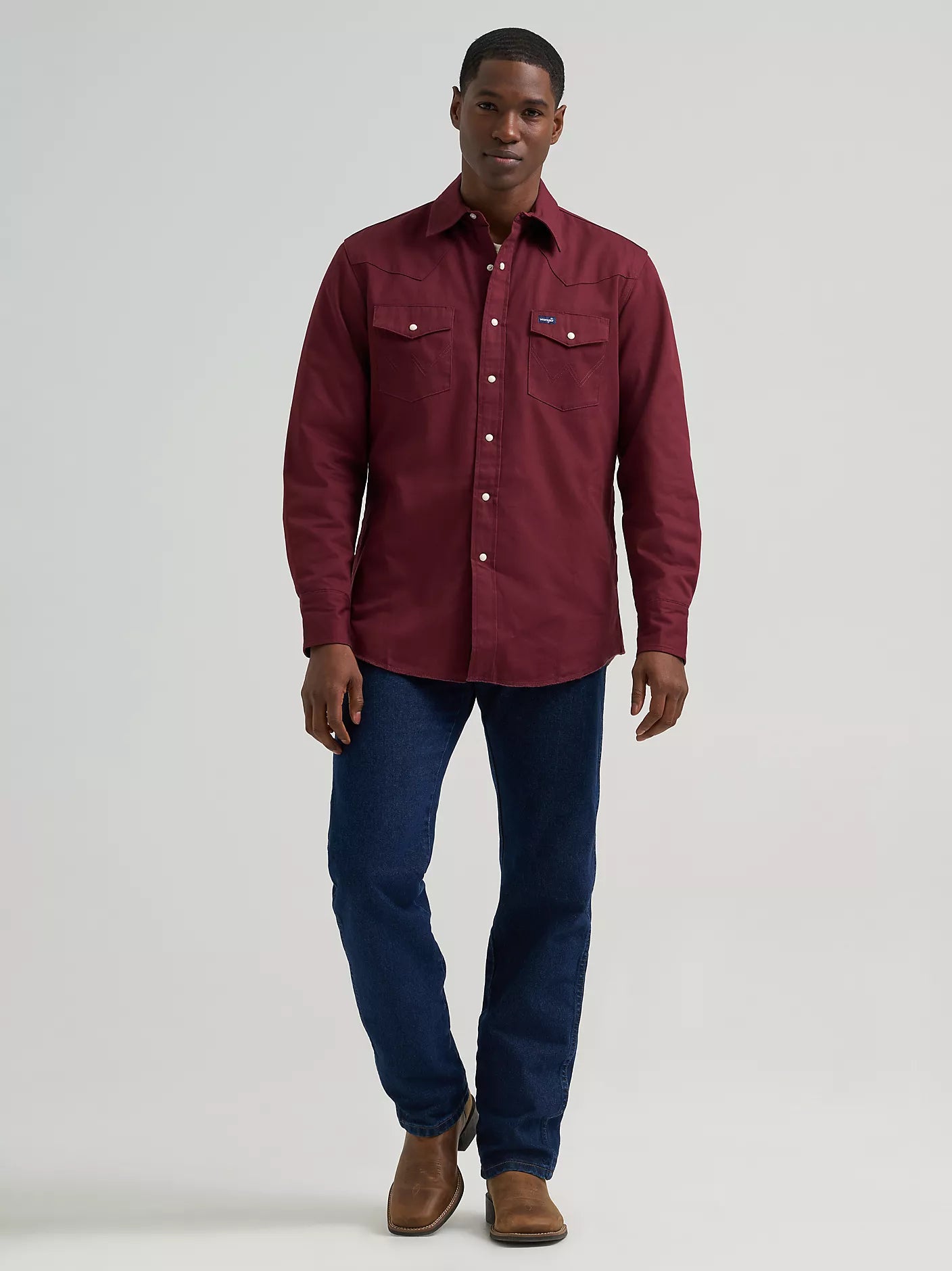 A man modeling a burgundy long-sleeve western-style button-up shirt with two chest pockets, paired with dark blue jeans and brown boots, against a light gray studio background.