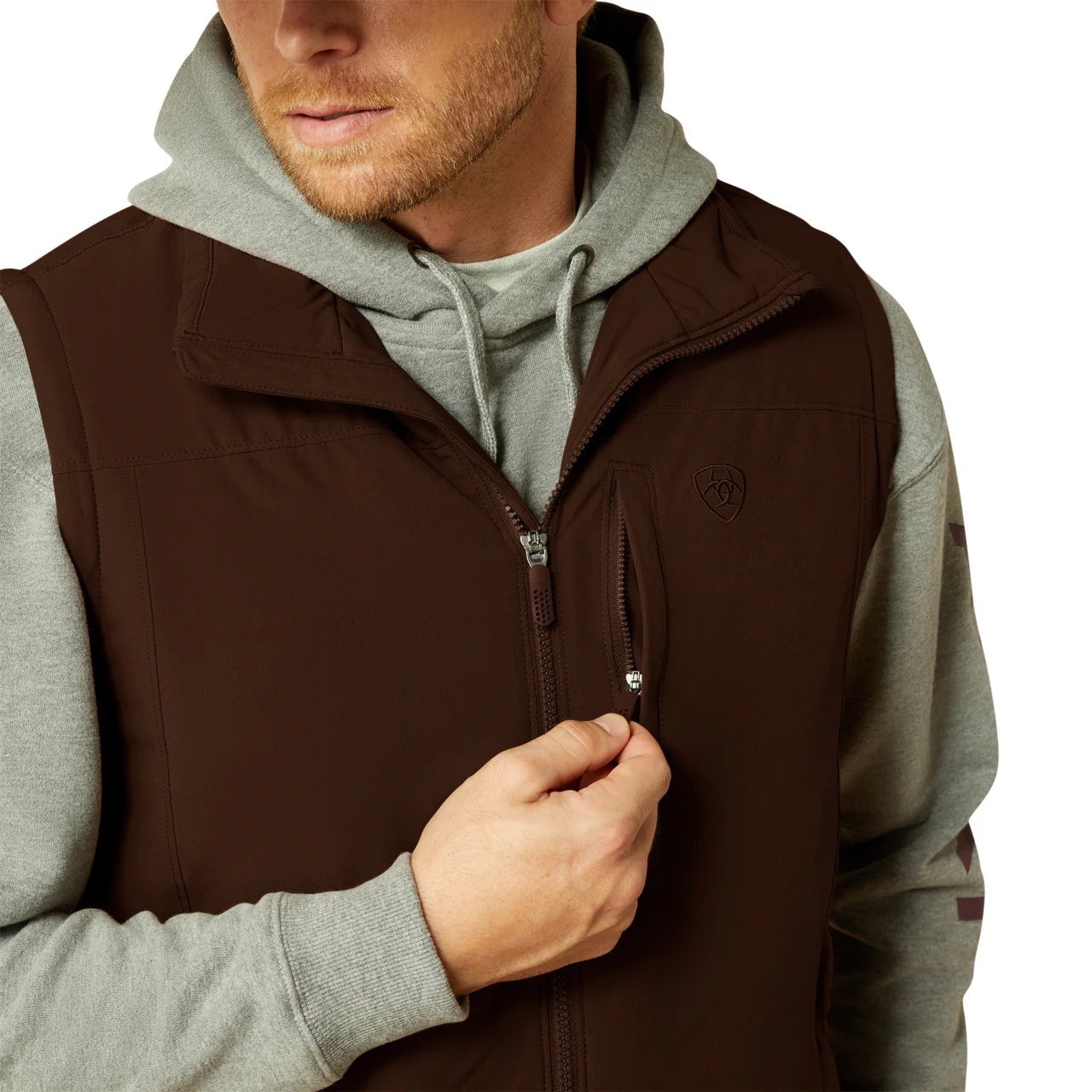 Man wearing brown zip-up vest over grey hoodie, using chest pocket, casual outdoor apparel