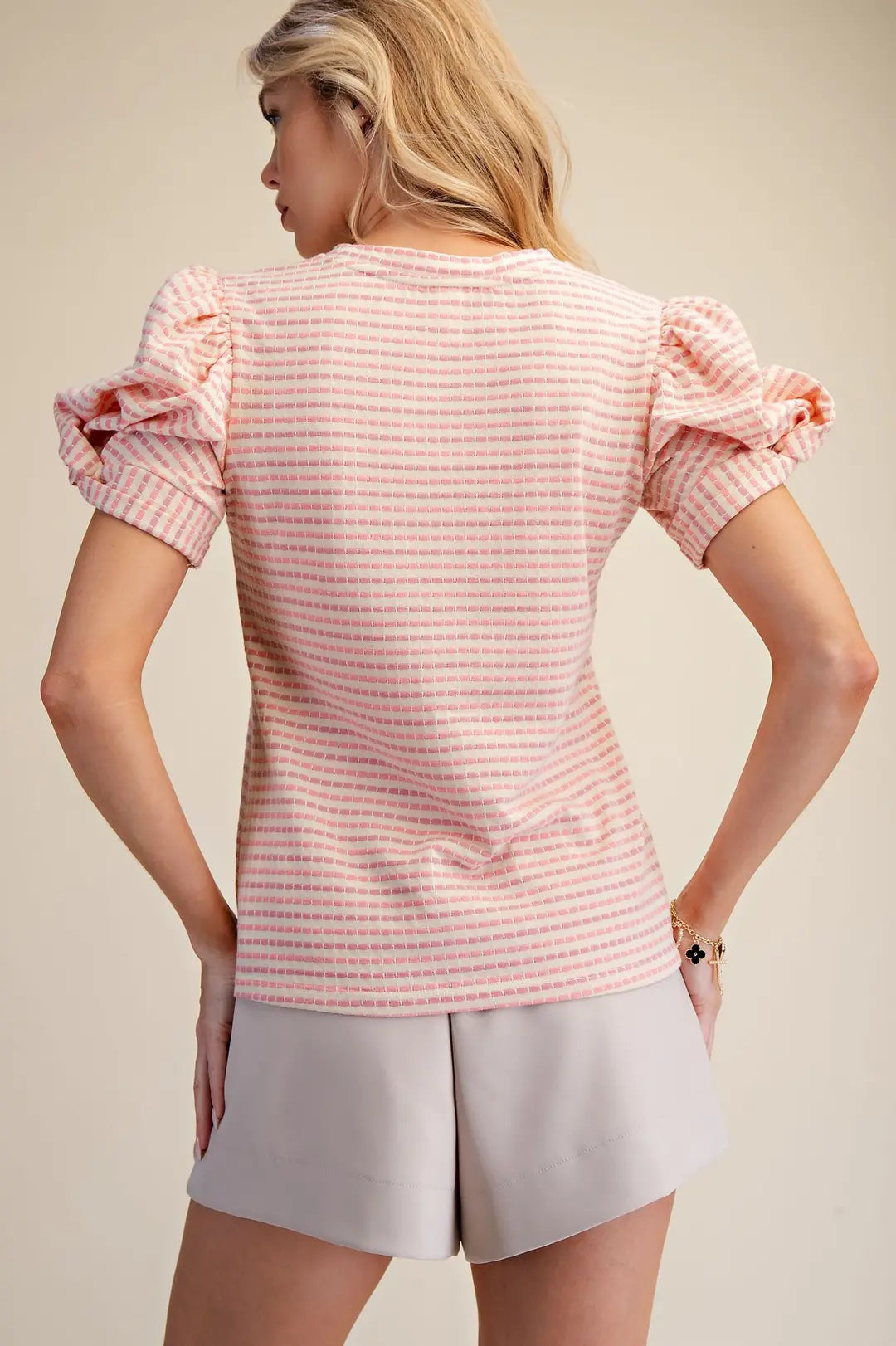 Woman wearing a pink and white striped shirt with ruffled sleeves and light gray shorts on a beige background