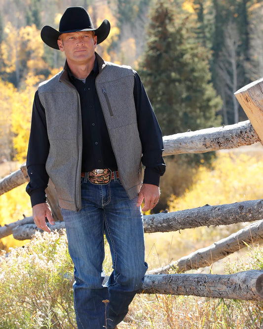 Man in cowboy hat, grey vest, and jeans standing by wooden fence in autumn countryside