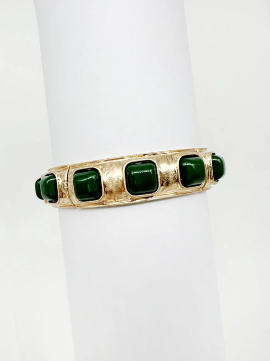 Gold cuff bracelet with square green stones, displayed on a white cylinder
