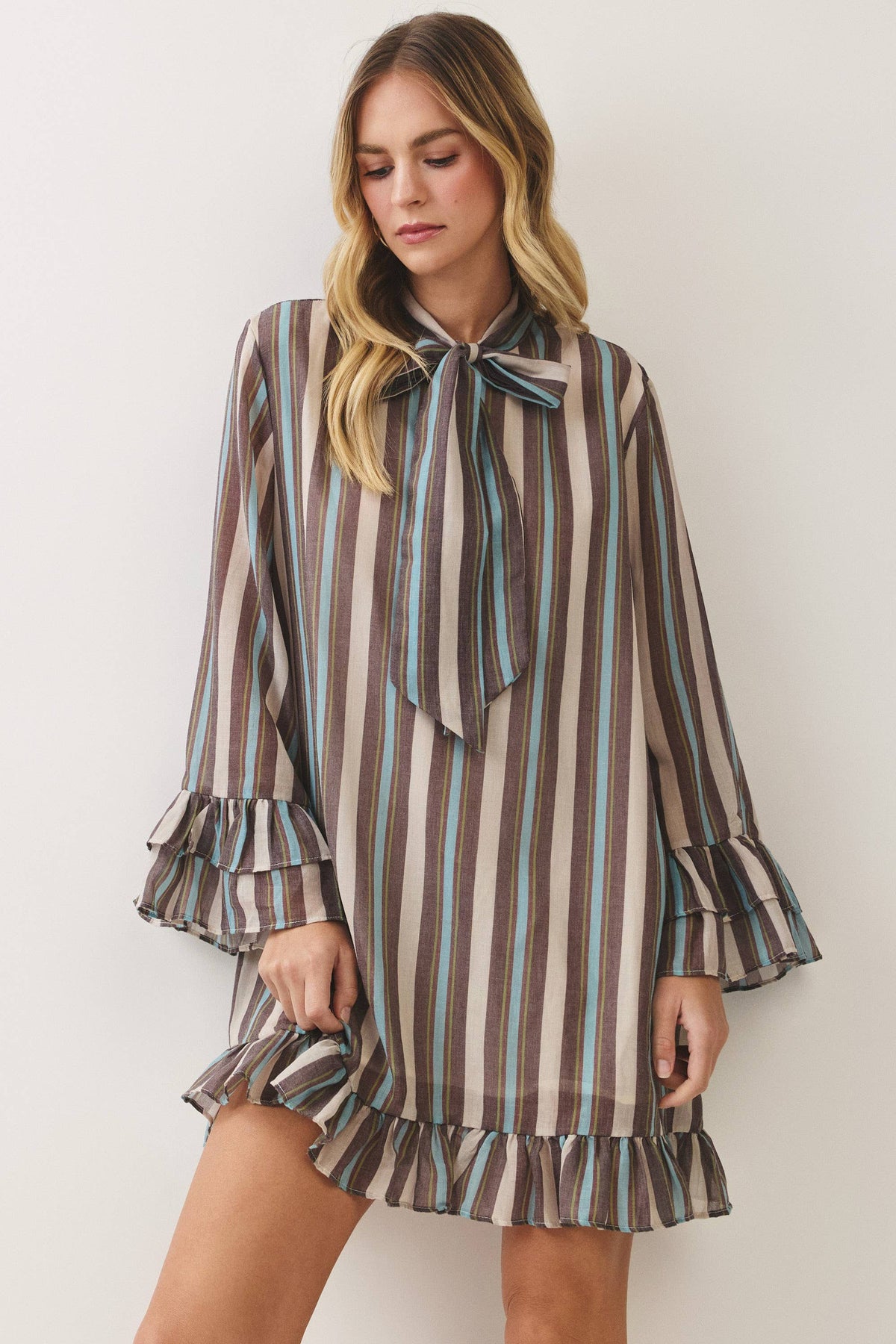 A woman wearing a brown, beige, and light blue vertically striped mini dress with a tie-neck bow and ruffled cuffs and hem, posing against a plain off-white background.