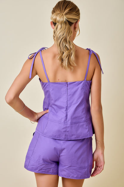A woman modeling a matching lavender purple two-piece set consisting of a tie-strap crop top and coordinating shorts, set against a solid beige background.