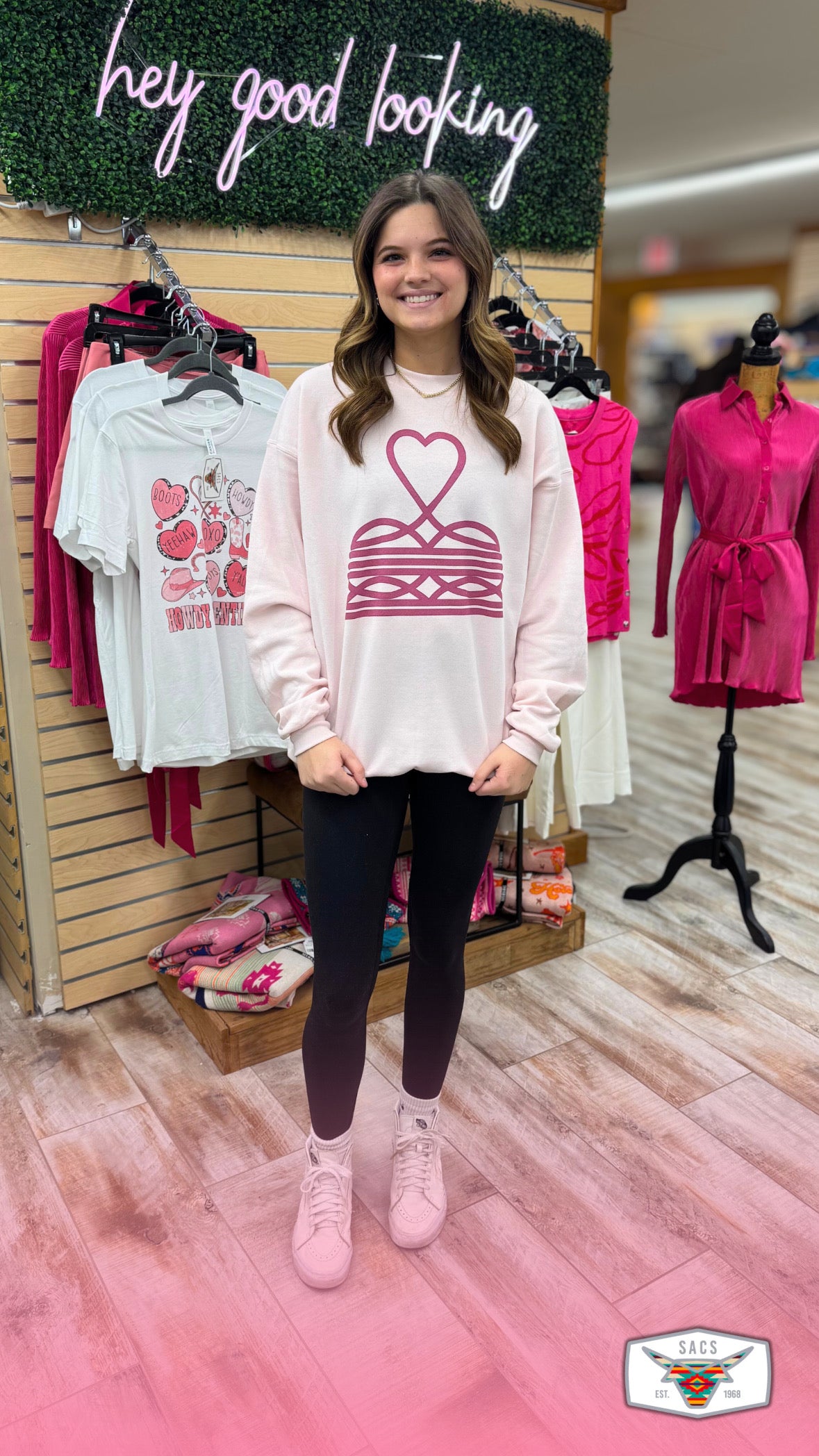 Smiling woman in casual pink graphic sweatshirt, black leggings, and sneakers standing in boutique.
