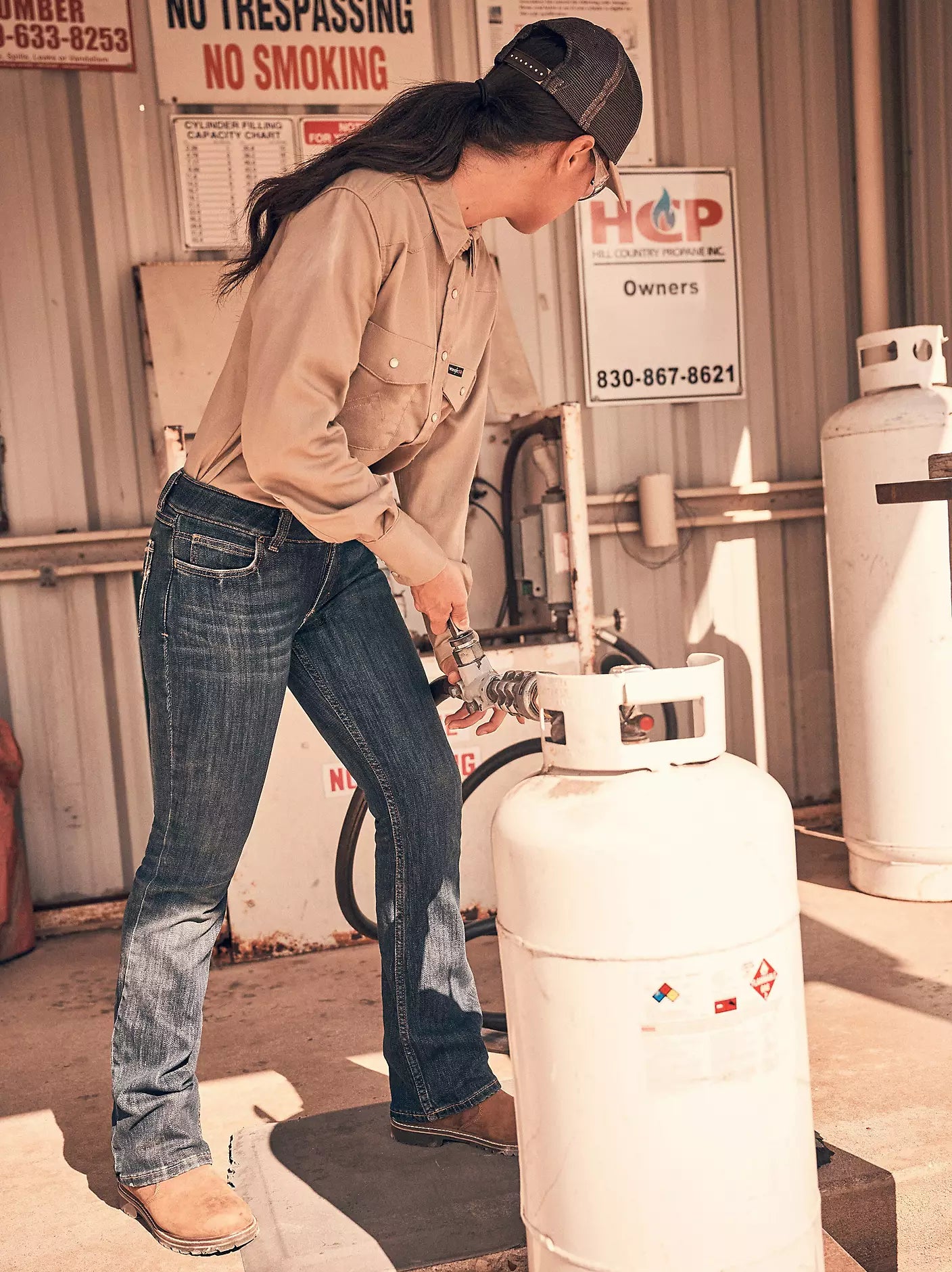 Person in work shirt, jeans, and boots filling propane tank outdoors at gas refill station