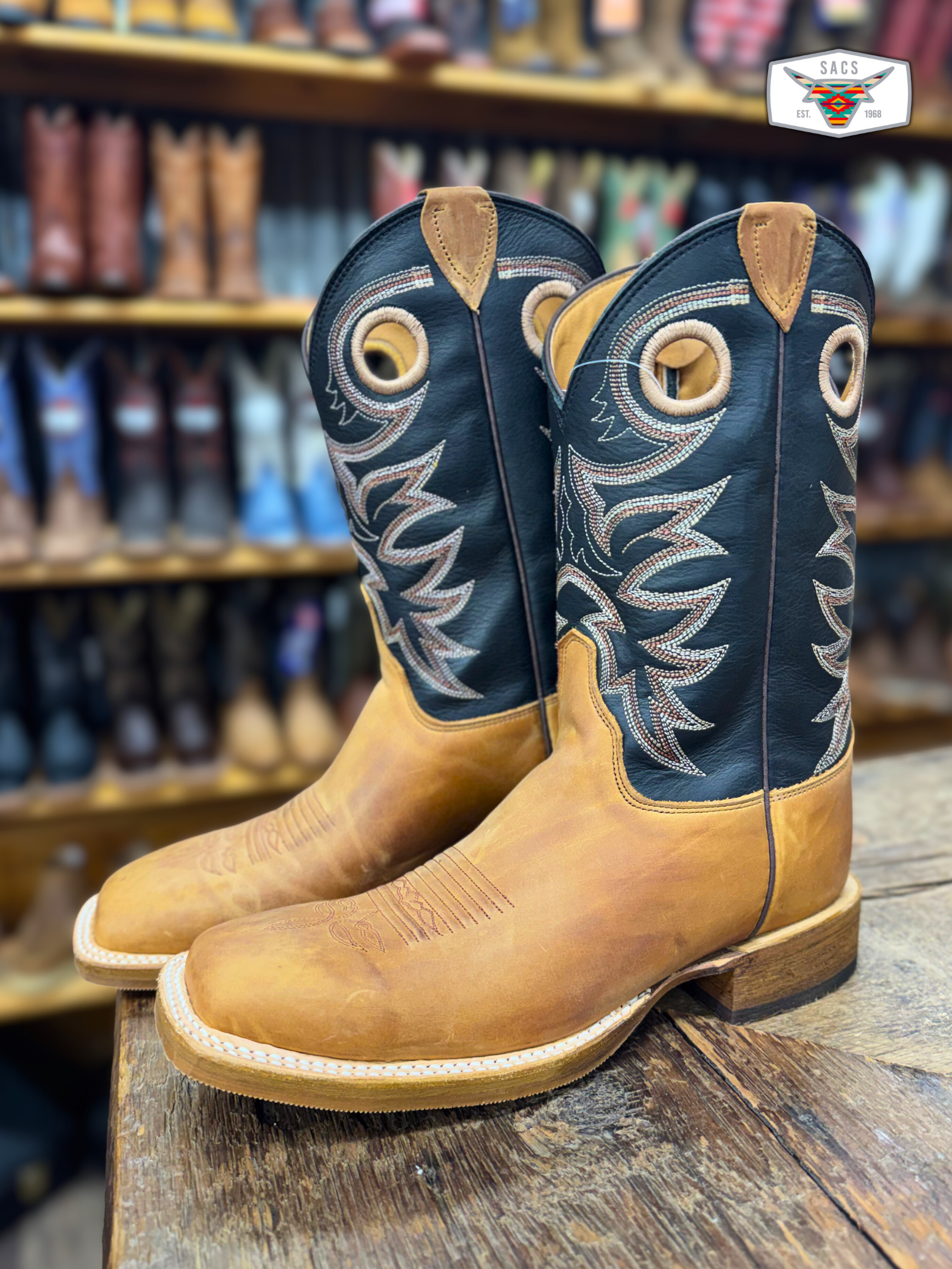 Brown leather cowboy boots with black embroidered shafts on a rustic wood table in a western store.