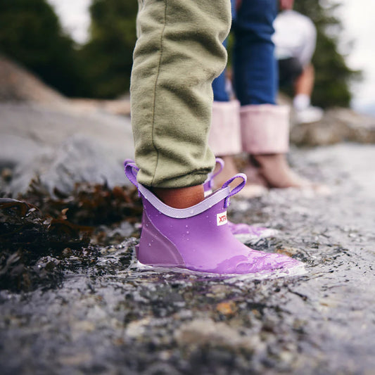 Purple rain boots worn by a child standing in a puddle with a natural background.