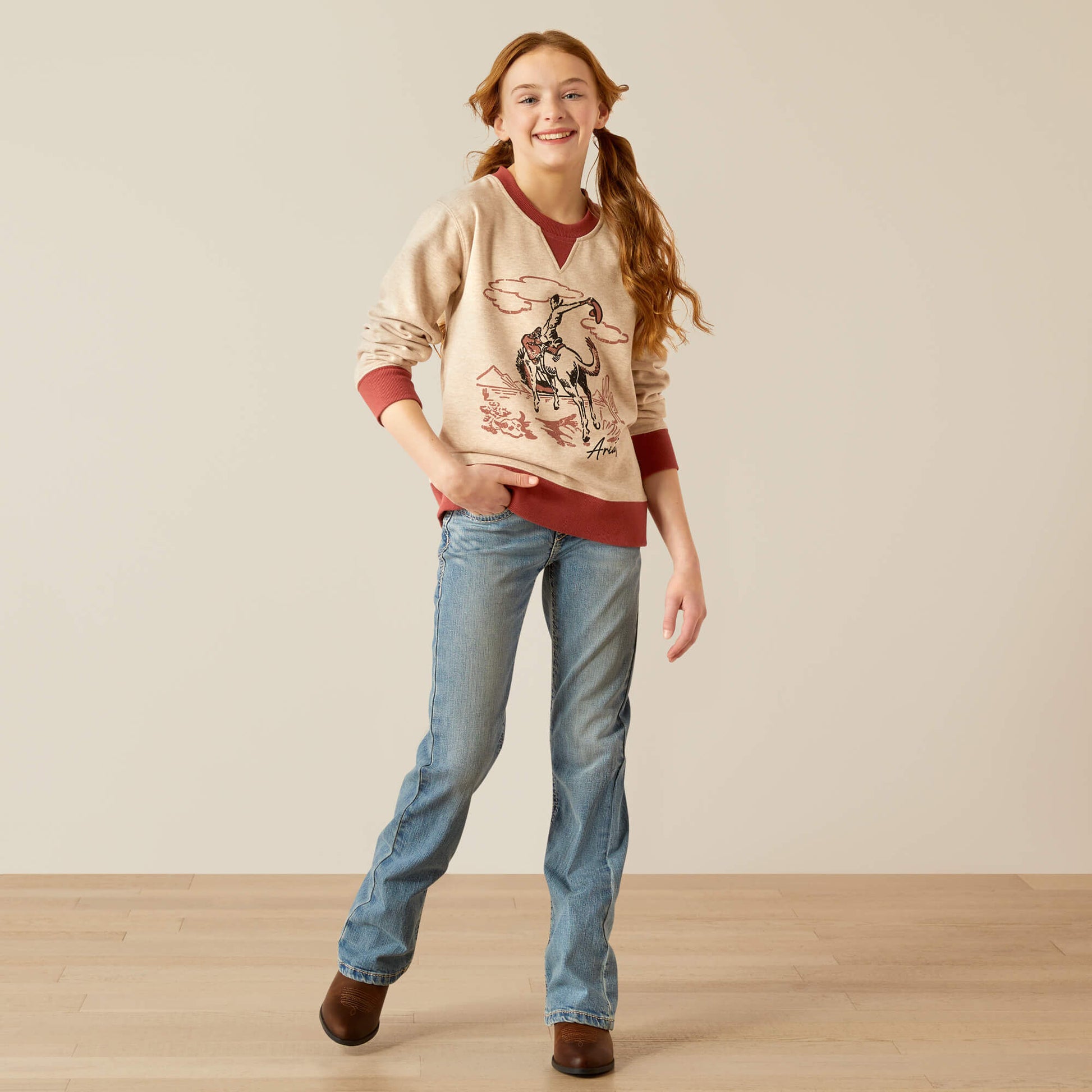 Smiling girl in western graphic sweatshirt, blue jeans, and brown boots on light wood floor.