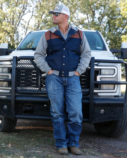 Man in outdoor workwear vest, hoodie, jeans, and cap standing in front of pickup truck