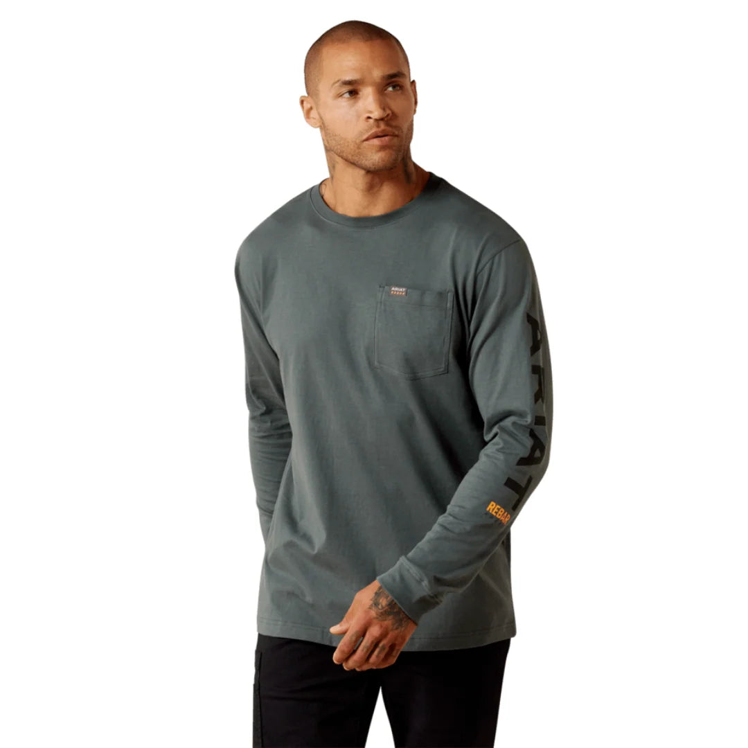 A man wearing a muted slate blue long-sleeve pocket shirt with black pants, standing against a plain beige studio background.