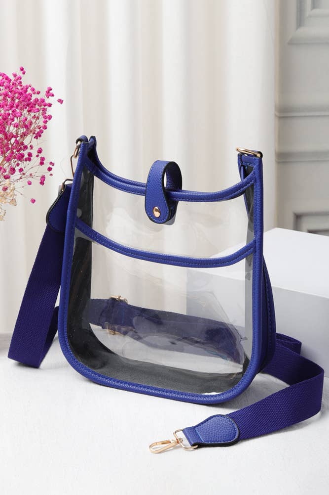 A clear plastic bucket-style shoulder bag with a vibrant blue trim, strap, and snap closure, displayed on a white surface next to pink flowers.