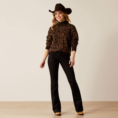 Woman wearing a brown patterned sweater and black pants with a cowboy hat on a plain background