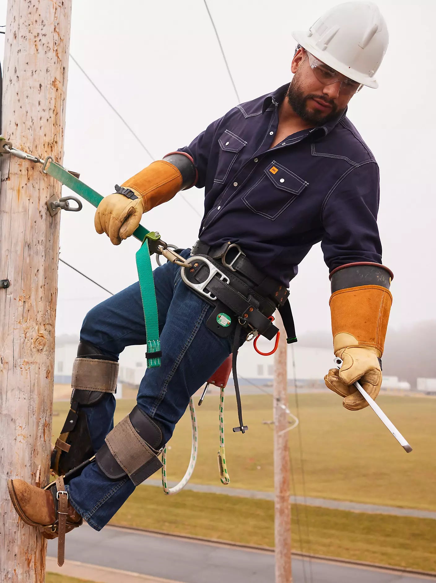 Utility worker in safety gear climbing a power pole with harness, gloves, and hard hat outdoors.