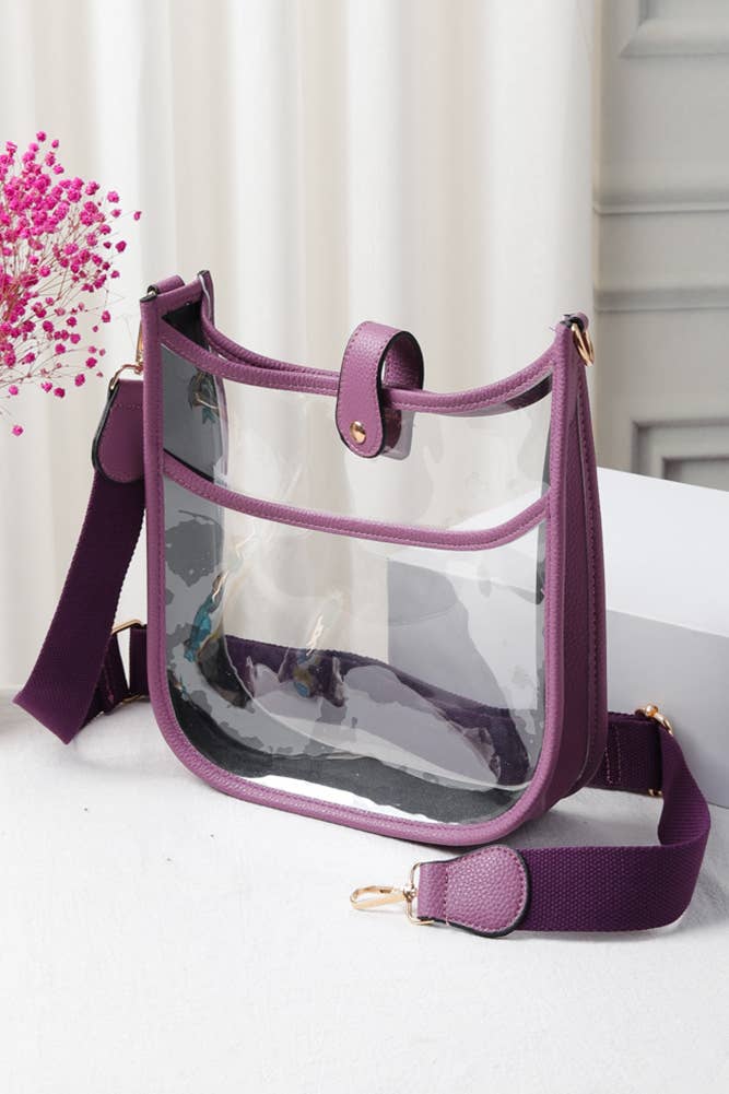 A clear plastic shoulder bag with a purple faux-leather trim and a matching purple adjustable webbing strap, displayed on a white surface with a decorative pink flower in the background.