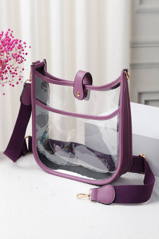 A clear plastic shoulder bag with a purple faux-leather trim and a matching purple adjustable webbing strap, displayed on a white surface with a decorative pink flower in the background.