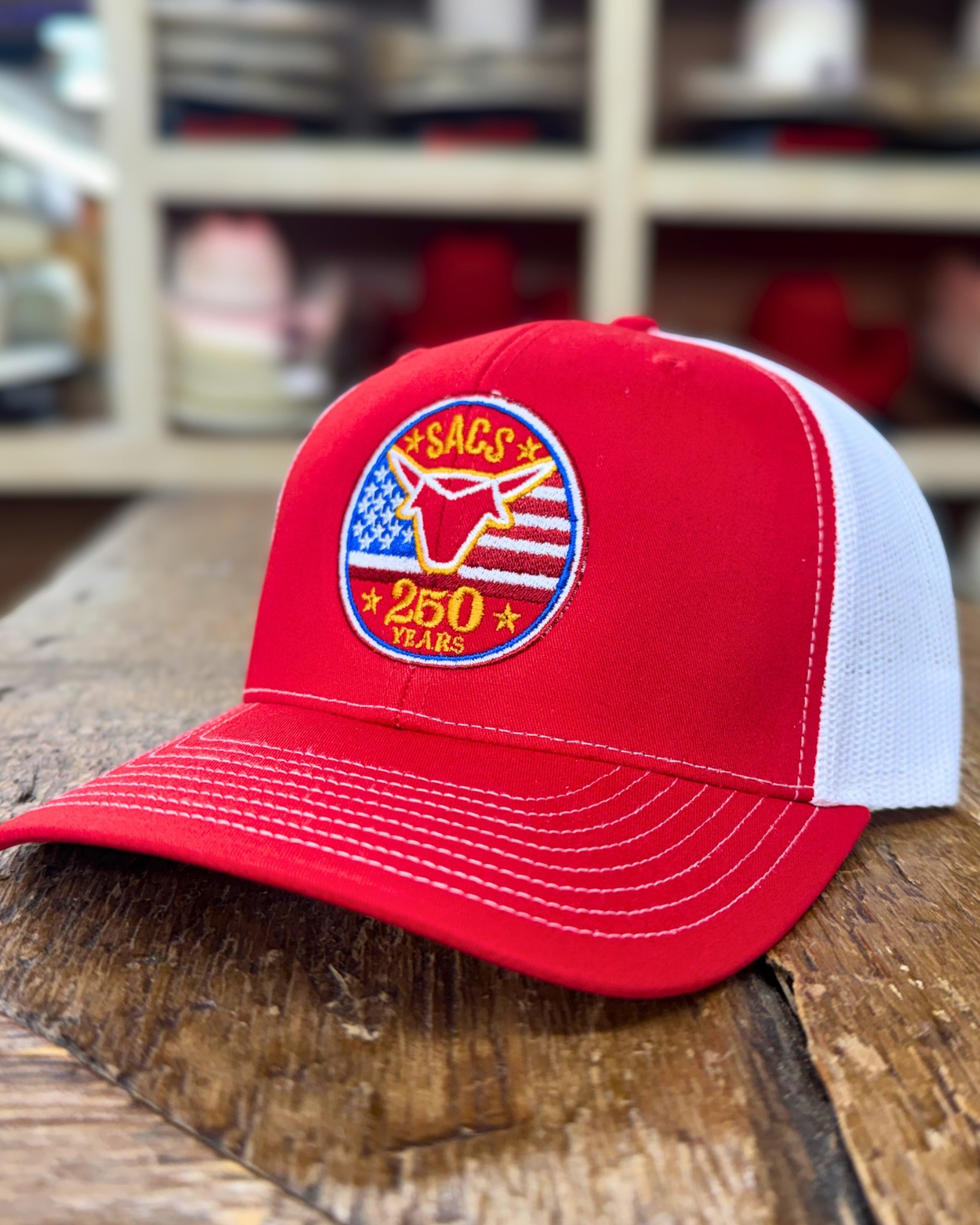 Red and white cap with a logo on a wooden surface, with shelves of caps in the background.