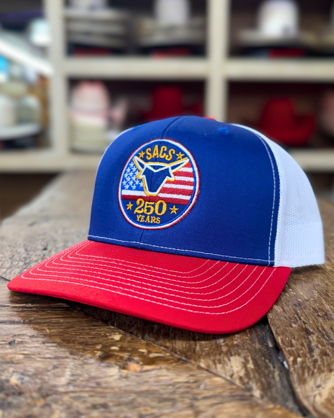 Blue and red cap with a logo on a wooden surface, with shelves of caps in the background.