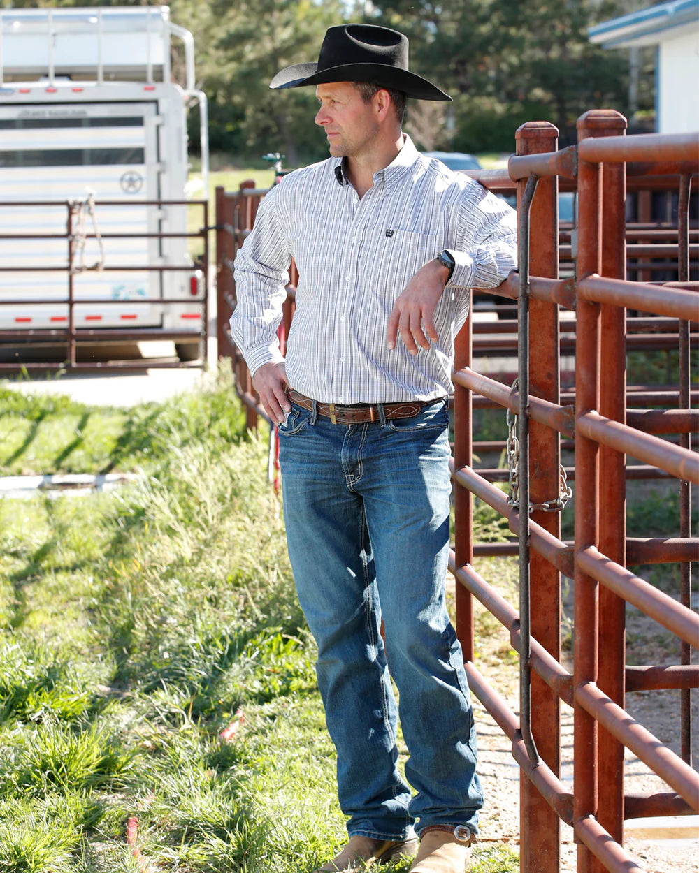 Man in cowboy hat, striped shirt, and jeans leaning on ranch fence outdoors