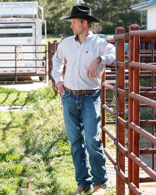 Man in cowboy hat, striped shirt, and jeans leaning on ranch fence outdoors