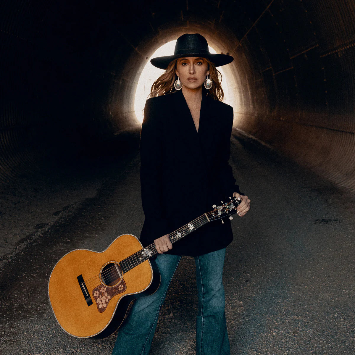 Lainey Wilson holding an acoustic guitar in a tunnel setting, wearing a black Charlie 1 Horse Hat