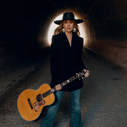 Lainey Wilson holding an acoustic guitar in a tunnel setting, wearing a black Charlie 1 Horse Hat