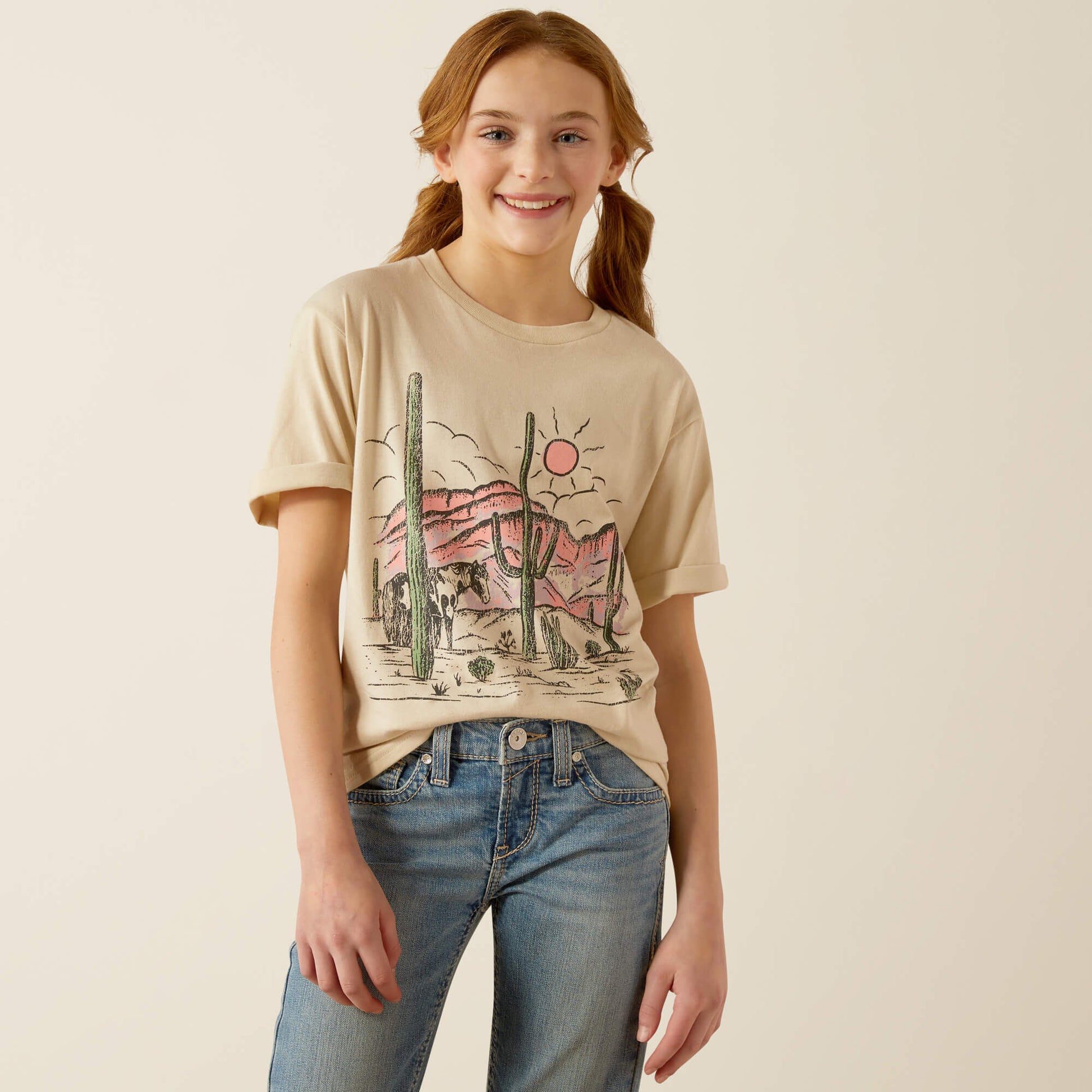 Smiling girl in a beige desert graphic t-shirt and blue jeans against neutral background
