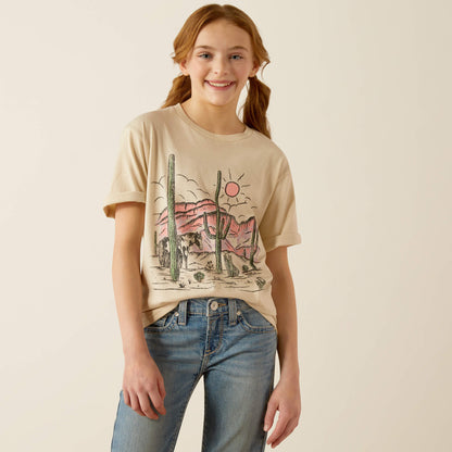 Smiling girl in a beige desert graphic t-shirt and blue jeans against neutral background