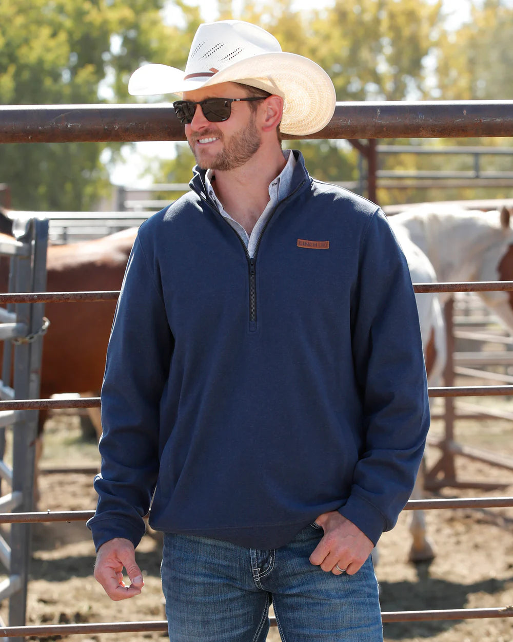 Man in navy quarter-zip sweater, cowboy hat, sunglasses, standing by horse pen outdoors